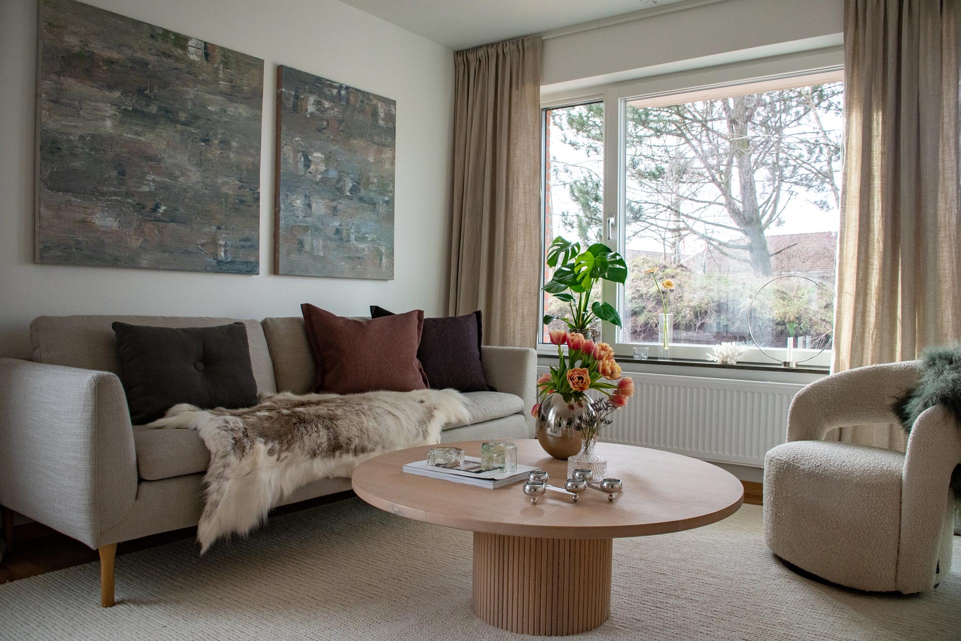 Ett välbelyst och mysigt vardagsrum med en neutral färgpalett. Ett stort fönster släpper in naturligt ljus som framhäver de inomhusväxter och buketten med blommor som finns på soffbordet. Möblerna inkluderar en beige soffa med kuddar, ett runt soffbord och en fåtölj.