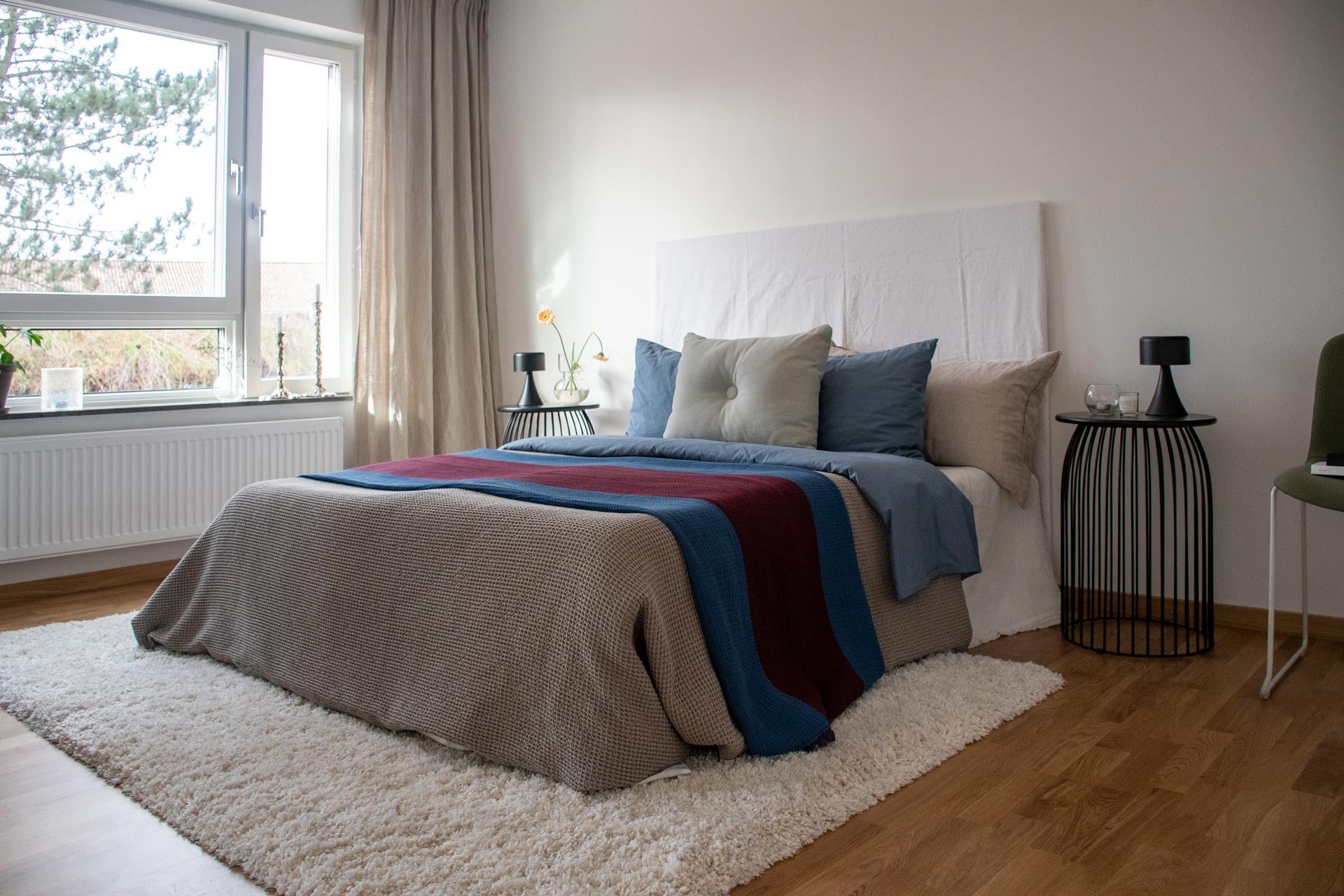 Ett prydligt sovrum med ett stort fönster som släpper in naturligt ljus. Sängen har en vit gavel och är täckt med ett grått täcke och en blå pläd. Flera kuddar är placerade mot gaveln. På golvet ligger en fluffig vit matta, och vid ena sidan av sängen finns ett litet nattduksbord med en lampa.